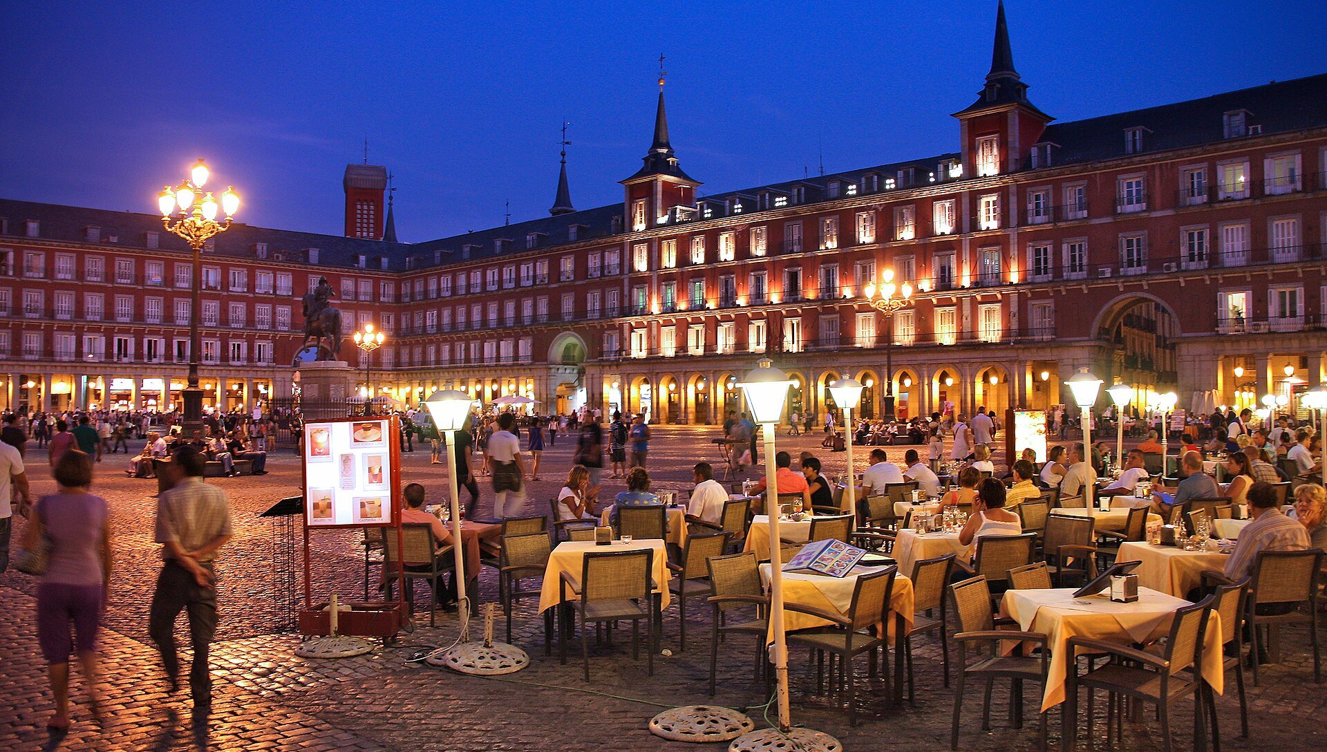 Plaza Mayor de Madri à noite, com mesas de restaurante ocupadas sob as arcadas iluminadas