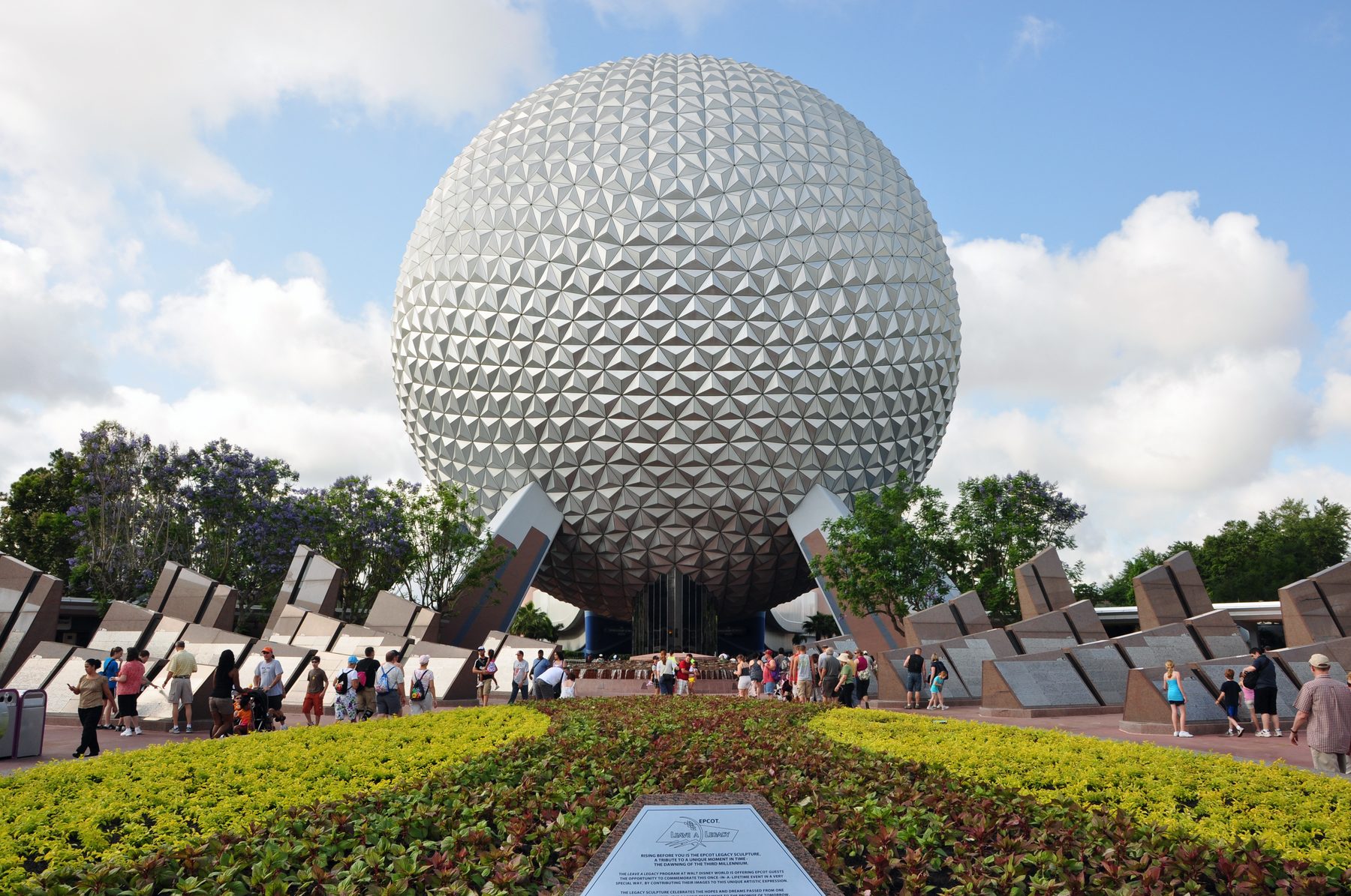 Esfera geodésica Spaceship Earth vista de frente sob céu de nuvens brancas, com canteiro florido e visitantes caminhando na entrada do EPCOT