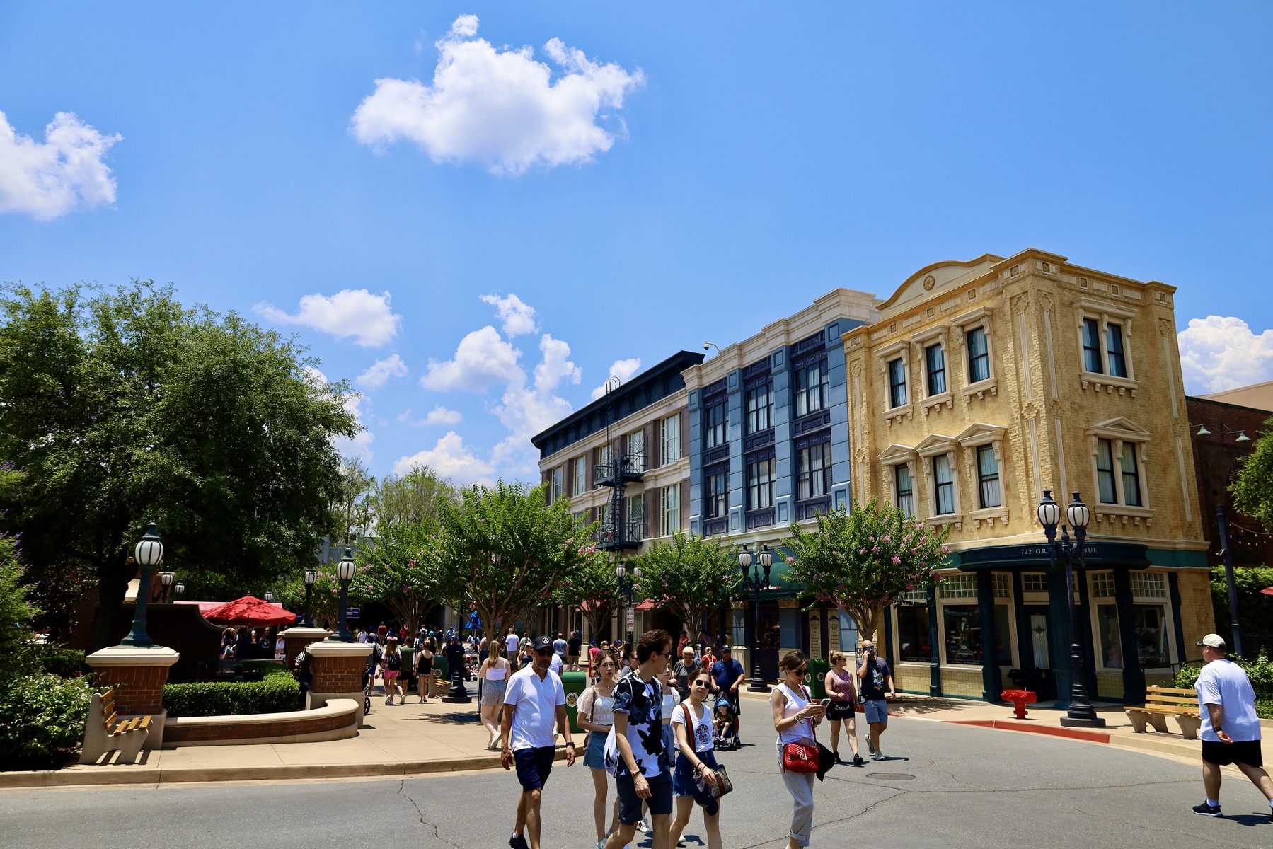 Fachadas em tons de creme e azul de Grand Avenue no Hollywood Studios sob céu azul com nuvens, com visitantes atravessando a rua