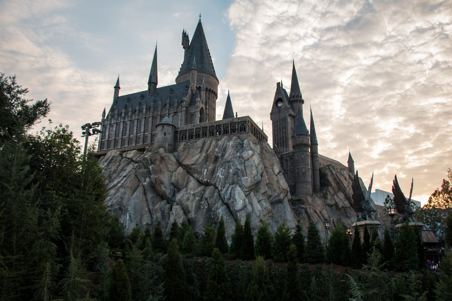 Castelo de Hogwarts recortado contra céu de nuvens douradas no fim da tarde, erguido sobre rocha cinzenta acima de uma fileira de pinheiros