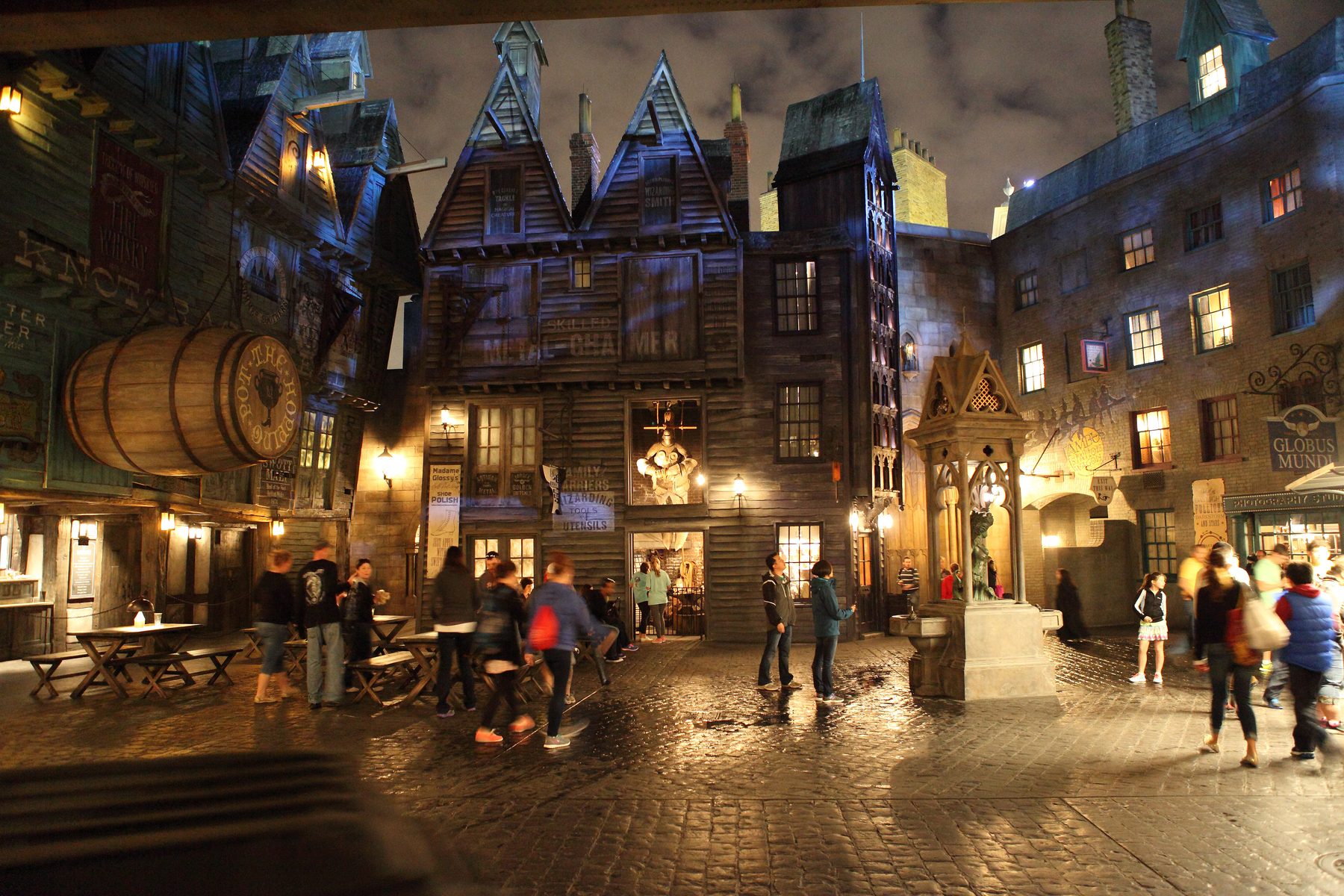 Diagon Alley à noite, com fachadas tortas de madeira iluminadas por lampiões e visitantes caminhando pelo calçamento molhado